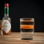 Un verre à shot contenant une couche de tequila transparente surmontée d'une fine bande de sauce Tabasco rouge vif, avec une bouteille de Tabasco en arrière-plan sur une surface en bois.