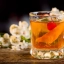 Verre à cocktail avec un mélange ambré, garni d'un zeste d'orange et d'une cerise rouge, posé sur une table en bois avec des fleurs blanches en arrière-plan.